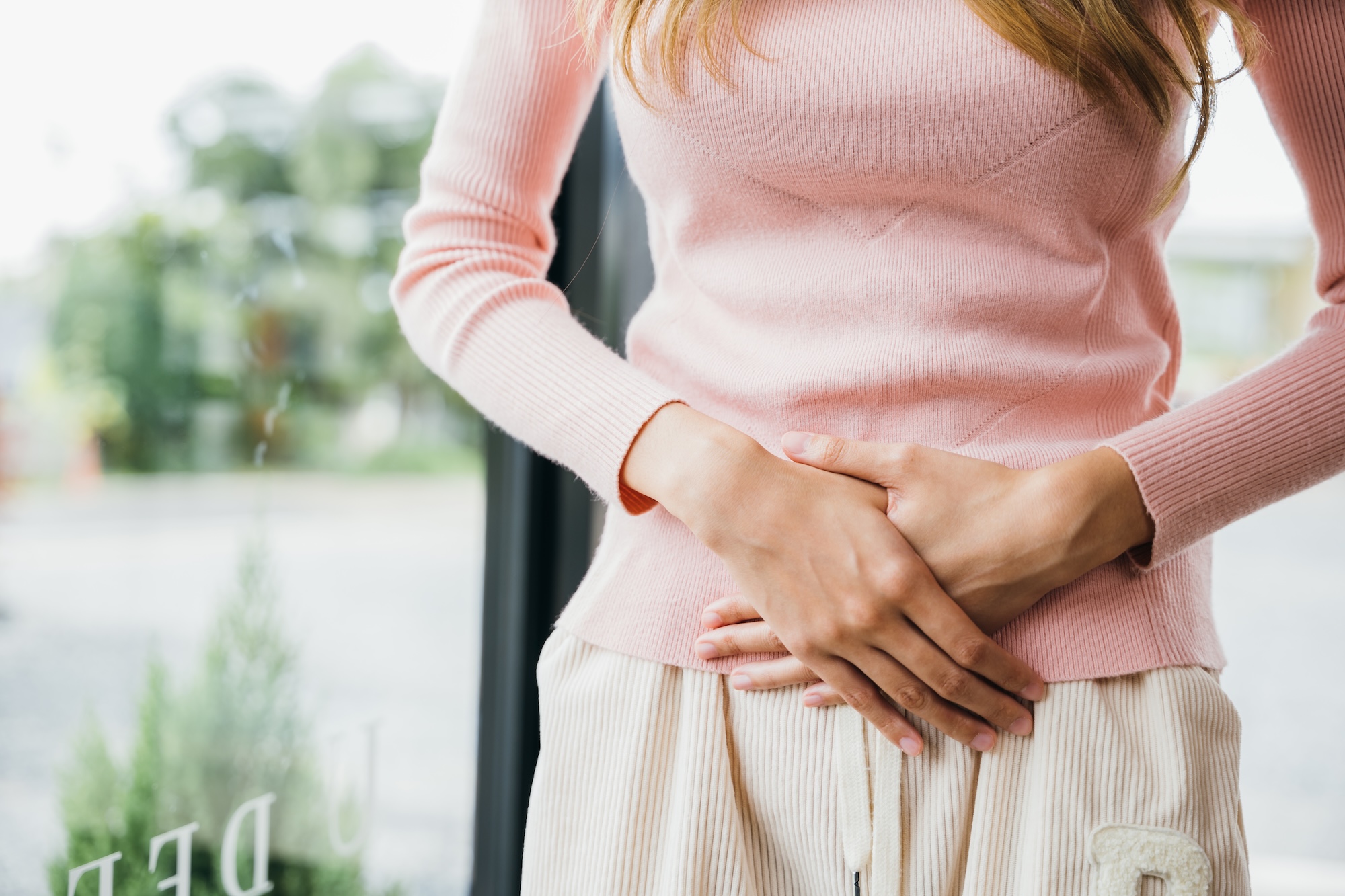 A woman at home in a pink sweater, holding her abdomen in discomfort from a dysfunctional pelvic floor. She can schedule an appointment with Harbour Direct Primary Care for pelvic floor dysfunction treatment near Newport.